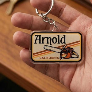May include: A rectangular keychain with the name "Arnold" in black, a chainsaw graphic, and the word "CALIFORNIA". The keychain has a clear border and a silver keyring.
