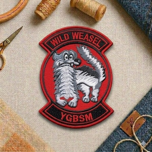 May include: Embroidered patch with the words "WILD WEASEL" in black on a red background. The patch features a cartoon weasel in gray and white. The text "YBGSM" is below the weasel. The patch is surrounded by black trim.