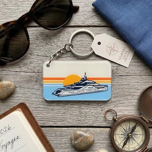 May include: A rectangular keychain featuring a yacht design with a sunset backdrop. The keychain has a white background with orange, yellow, and blue stripes. The yacht is white and navy blue. The keychain is attached to a silver chain and ring.