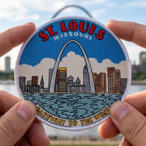 May include: A circular embroidered patch featuring the St. Louis, Missouri skyline with the Gateway Arch in the center. The text reads ST. LOUIS MISSOURI at the top and GATEWAY TO THE WEST at the bottom, set against a blue sky and water background.