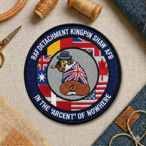 May include: A circular patch with a blue border and the text "RAF DETACHMENT KINGPIN SHAW AFB" and "IN THE 'ARCENT' OF NOWHERE". The patch features a cartoon dog wearing a Union Jack coat and a hat, surrounded by flags.