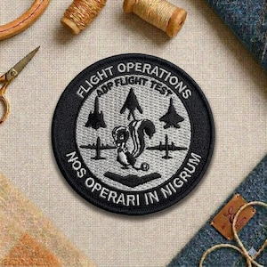 May include: A round, embroidered patch with the text "FLIGHT OPERATIONS ADP FLIGHT TEST" and "NOS OPERARI IN NIGRUM". The patch features a squirrel, airplanes, and a black border. The patch is on a beige fabric.