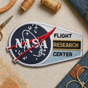 May include: Embroidered NASA patch featuring the NASA logo in white on a navy blue background, with a red swoosh. The patch also includes the text "FLIGHT RESEARCH CENTER" in white and gold lettering. The patch is surrounded by a white border.