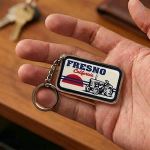 May include: A rectangular keychain featuring the text "FRESNO California" above a graphic of a tractor. The design includes a red and blue sunset. The keychain is attached to a silver chain and ring.