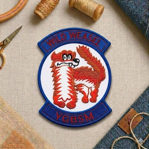 May include: Embroidered patch featuring a cartoon weasel in red and white, with the text "WILD WEASEL" in blue. The patch also includes the text "YCBSM" in red on a blue banner. The patch is circular and has a white background.