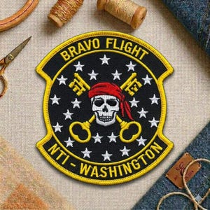 May include: Embroidered patch with a black background, yellow border, and white stars. The patch features the text "BRAVO FLIGHT" and "NTI - WASHINGTON". A skull wearing a red bandana and holding two gold keys is in the center.