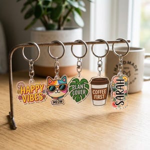 May include: A collection of acrylic keychains with various designs, including a cat wearing sunglasses, a coffee cup, a monstera leaf, and the words "Happy Vibes", "Plant Lover", and "Coffee First". Each keychain is attached to a silver keyring.