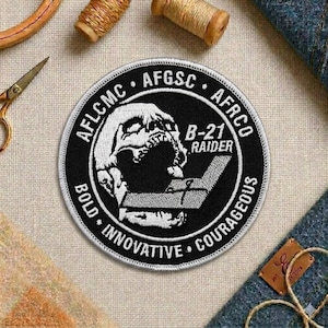May include: A round black and white embroidered patch featuring a stylized skull and the text "B-21 RAIDER". The patch also includes the words "BOLD", "INNOVATIVE", and "COURAGEOUS" around the edge, along with other text.