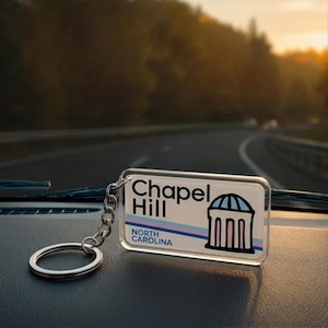 May include: A rectangular keychain with the text "Chapel Hill NORTH CAROLINA" in black and blue. The keychain features a graphic of a building with a dome. The background shows a blurred road and trees.