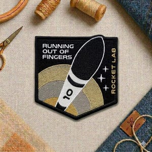 May include: Black and white embroidered patch with the text "RUNNING OUT OF FINGERS" and "ROCKET LAB". The patch features a black and white rocket design with the number 10. The patch is surrounded by gold and gray concentric circles.