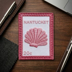 May include: A rectangular embroidered patch featuring a white center with a pink seashell design, the word NANTUCKET in pink text at the top, and the number 20c in pink text at the bottom, all framed by a thick pink stitched border.