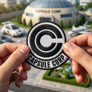 May include: A round, black and white embroidered patch with the text "CAPSULE CORP." and a stylized "C" logo. The patch is held up in front of a building with a dome-shaped roof and a small car.