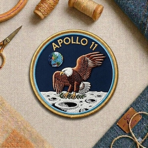 May include: Embroidered patch featuring the Apollo 11 mission. The patch has a blue background with a gold border, an eagle, a globe, and the words "Apollo 11". The eagle is brown and white, and the globe shows the Earth.