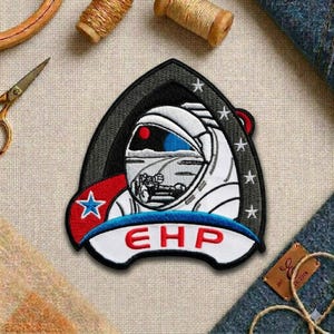May include: Embroidered patch featuring a futuristic astronaut helmet design with a red eye and blue visor. The patch includes the letters "EHP" and a star on a red and white banner. The patch is surrounded by a black and gray border and white stars.