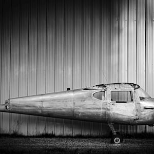May include: A black and white photo of a vintage airplane fuselage, partially visible, parked in front of a corrugated metal wall.