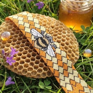 May include: A beaded bracelet with a bee motif, displayed on a honeycomb. The bracelet has a repeating pattern of orange and cream beads, with a detailed bee in the center. A small jar of honey and purple flowers are in the background.