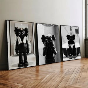 May include: Three framed black and white art prints depicting a cartoon character in bathroom scenes. The character has large ears, crossed-out eyes, and a white towel. The prints are displayed on a wooden floor.