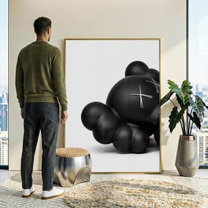May include: A large rectangular wall art piece featuring a black, rounded, cartoon-style character with X-shaped eyes on a white background, framed in a thin gold border. The artwork is displayed on a wall next to a metallic stool and a potted plant.