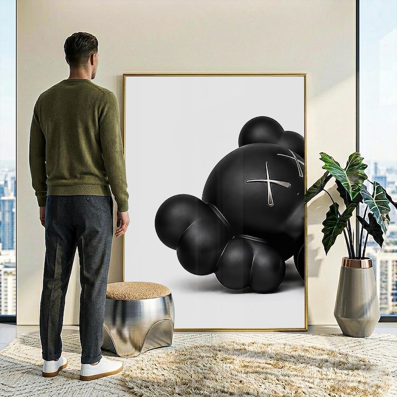 May include: A large rectangular wall art piece featuring a black, rounded, cartoon-style character with X-shaped eyes on a white background, framed in a thin gold border. The artwork is displayed on a wall next to a metallic stool and a potted plant.