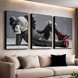 Pode incluir: Tríptico de arte de parede com uma figura estilizada em uma pose relaxada. A obra de arte é principalmente cinza e preta, com luvas brancas e um rosto branco com olhos riscados. A figura usa sapatos pretos com detalhes em vermelho. A arte é emoldurada em preto.