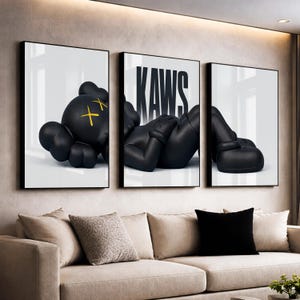 Pode incluir: Um tríptico de impressões artísticas emolduradas com uma figura preta em uma pose relaxada. A impressão central exibe a palavra "KAWS" em letras pretas em negrito. A figura tem olhos amarelos em forma de cruz. A obra de arte é exibida acima de um sofá bege.
