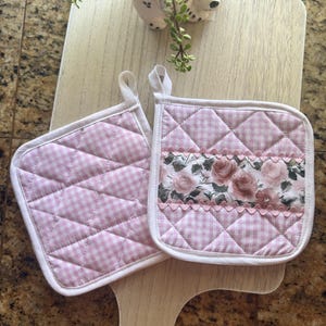 May include: Two square quilted pot holders featuring a light pink and white gingham pattern. One pot holder includes a central horizontal band decorated with a floral rose print and pink scalloped trim. Both items have white fabric hanging loops.