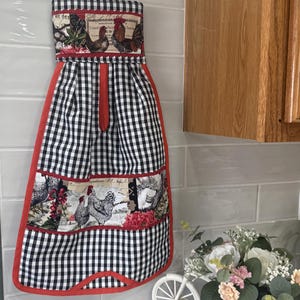 May include: A hanging kitchen towel featuring a black and white gingham pattern with red trim. The towel includes two horizontal fabric panels decorated with illustrations of roosters, chickens, and vintage-style French script text.