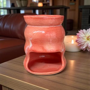 Może przedstawiać: Koralowy ceramiczny podgrzewacz do olejków o unikalnym, organicznym kształcie. Podgrzewacz ma duży otwór z przodu i szeroki wierzch. Powierzchnia ma błyszczące wykończenie z subtelnym białym marmurkiem. Stoi na drewnianym stole, z zapaloną świecą i kwiatem w tle.