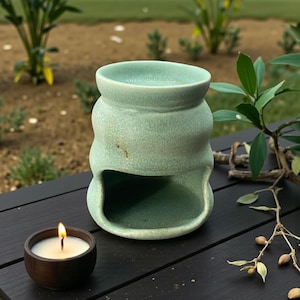 May include: A pale green ceramic oil diffuser with a unique, curved shape and an opening at the bottom. A small, round, wooden candle holder with a lit candle is placed beside the diffuser. The items are on a dark surface with greenery in the background.
