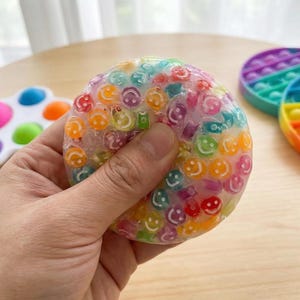 Op de afbeelding: Een doorzichtige, zachte stressbal gevuld met kleurrijke smiley-gezichten. De bal wordt in een hand gehouden en lijkt gedeeltelijk te zijn ingedrukt. Andere kleurrijke fidget-speeltjes zijn zichtbaar op de achtergrond.