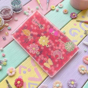 May include: A rectangular, clear resin piece filled with red beads and decorative charms, including a guitar, leaf, bow, flower, butterfly, and crescent moon. The background features colorful wooden planks and craft supplies.