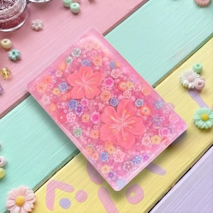 Peut inclure: Un étui de téléphone rectangulaire en résine rose avec un motif floral. L'étui présente deux grandes fleurs de cerisier roses et de nombreuses petites fleurs de différentes couleurs. L'arrière-plan est rempli de minuscules perles colorées.