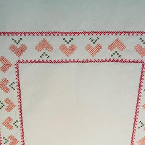 Puede incluir: Tela blanca con un borde bordado en rojo y rosa. El borde presenta un patr&oacute;n repetido de corazones rosas y detalles verdes. Los bordes est&aacute;n acabados con un punto de cruz rojo.