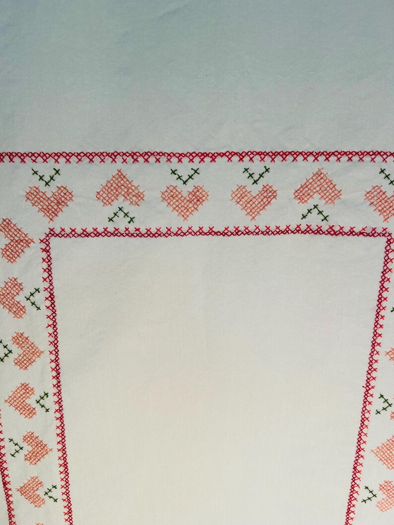 Puede incluir: Tela blanca con un borde bordado en rojo y rosa. El borde presenta un patr&oacute;n repetido de corazones rosas y detalles verdes. Los bordes est&aacute;n acabados con un punto de cruz rojo.