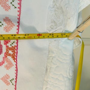 Puede incluir: Tela blanca con bordado de punto de cruz en rosa, naranja y verde. Una cinta m&eacute;trica amarilla muestra un ancho de aproximadamente 127 cm. La tela tiene un ribete de encaje y est&aacute; sujeta con una pinza de madera.