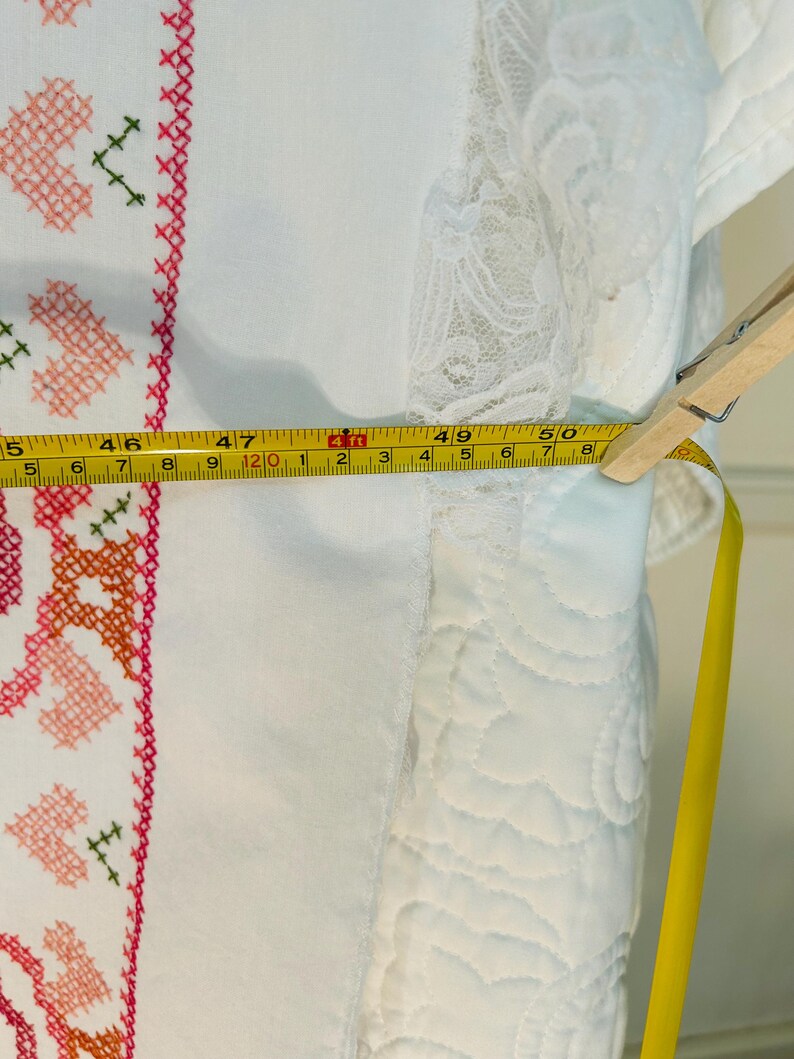 Puede incluir: Tela blanca con bordado de punto de cruz en rosa, naranja y verde. Una cinta m&eacute;trica amarilla muestra un ancho de aproximadamente 127 cm. La tela tiene un ribete de encaje y est&aacute; sujeta con una pinza de madera.