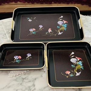 May include: A set of three rectangular black serving trays with gold-toned edges and integrated handles wrapped in light-colored cord. Each tray features a central dark brown panel with a woven texture and a floral design of morning glory flowers.