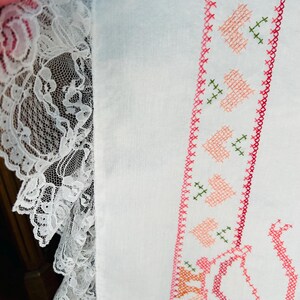 Puede incluir: Primer plano de tela blanca con un borde de punto de cruz detallado con corazones rosas y detalles verdes. Un delicado borde de encaje blanco a&ntilde;ade un toque vintage. El dise&ntilde;o sugiere un art&iacute;culo hecho a mano.