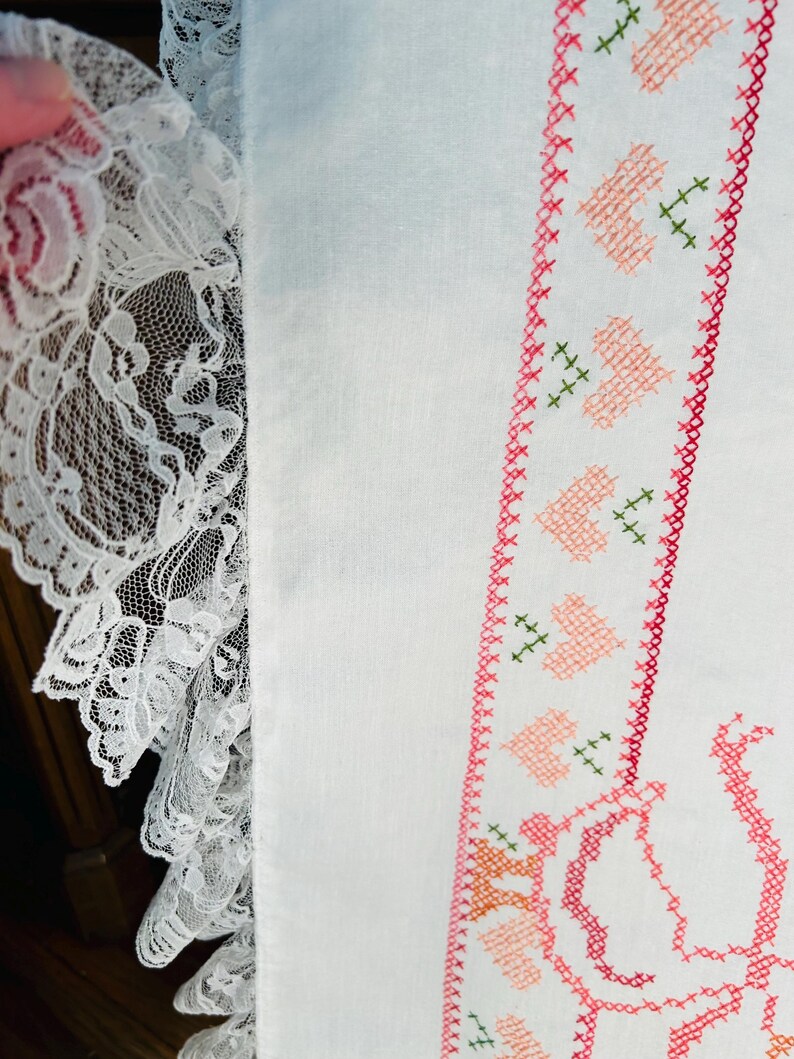 Puede incluir: Primer plano de tela blanca con un borde de punto de cruz detallado con corazones rosas y detalles verdes. Un delicado borde de encaje blanco a&ntilde;ade un toque vintage. El dise&ntilde;o sugiere un art&iacute;culo hecho a mano.