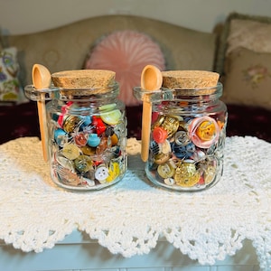 May include: Two clear glass jars with cork lids, each featuring a small wooden spoon attached to the side. The jars are filled with a variety of colorful, multi-shaped buttons in shades of red, blue, yellow, gold, and clear, showcasing diverse textures.
