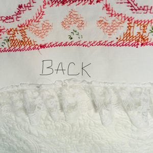 Puede incluir: Textil blanco con detalles bordados en rojo, naranja y verde. La palabra "BACK" est&aacute; escrita a mano en negro. En la parte inferior se ve un ribete de encaje blanco, con un patr&oacute;n acolchado debajo.
