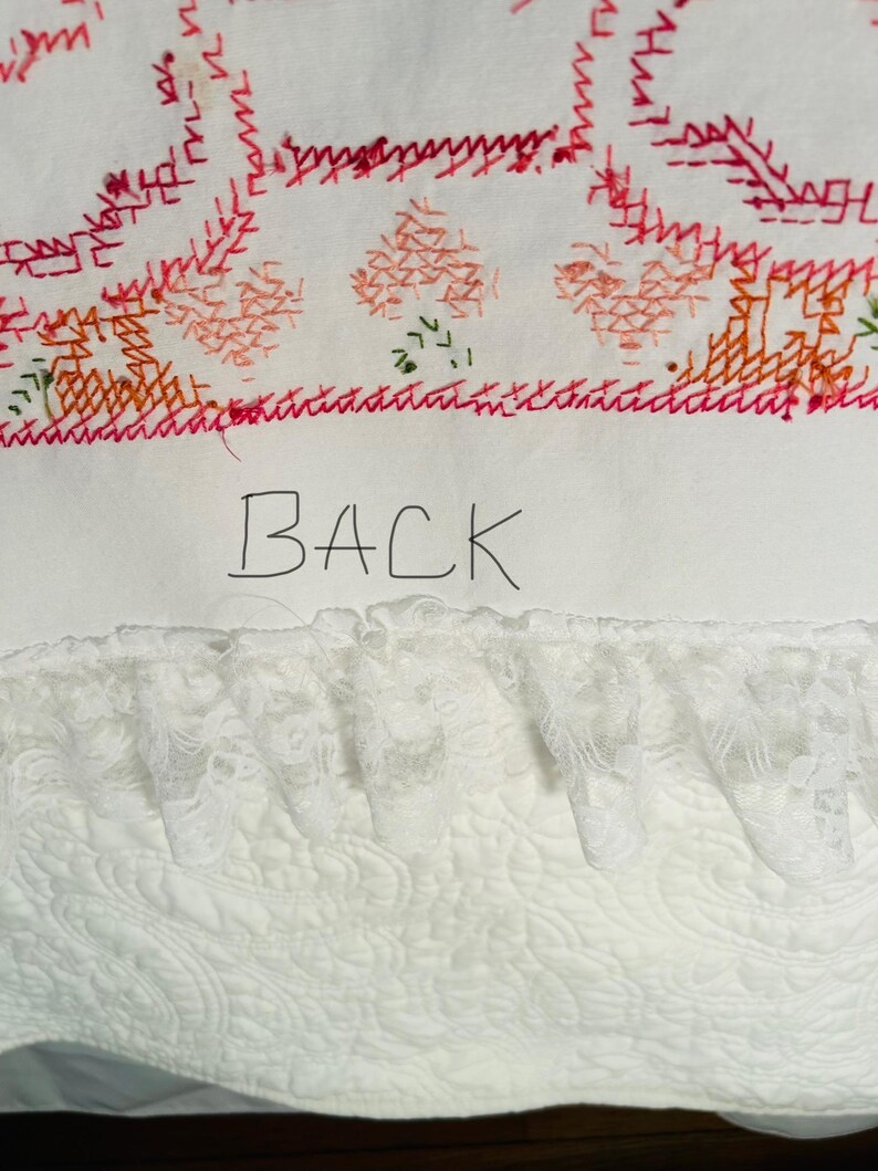 Puede incluir: Textil blanco con detalles bordados en rojo, naranja y verde. La palabra "BACK" est&aacute; escrita a mano en negro. En la parte inferior se ve un ribete de encaje blanco, con un patr&oacute;n acolchado debajo.