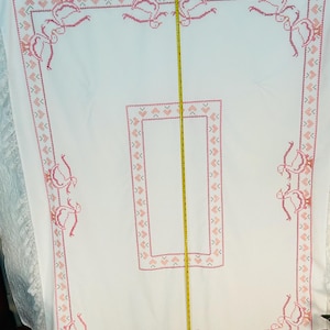 Puede incluir: Mantel blanco con un dise&ntilde;o de punto de cruz rosa. El borde presenta un patr&oacute;n repetido de corazones y p&aacute;jaros estilizados. El mantel tiene un ribete de encaje en el borde inferior. Se ve una cinta m&eacute;trica.