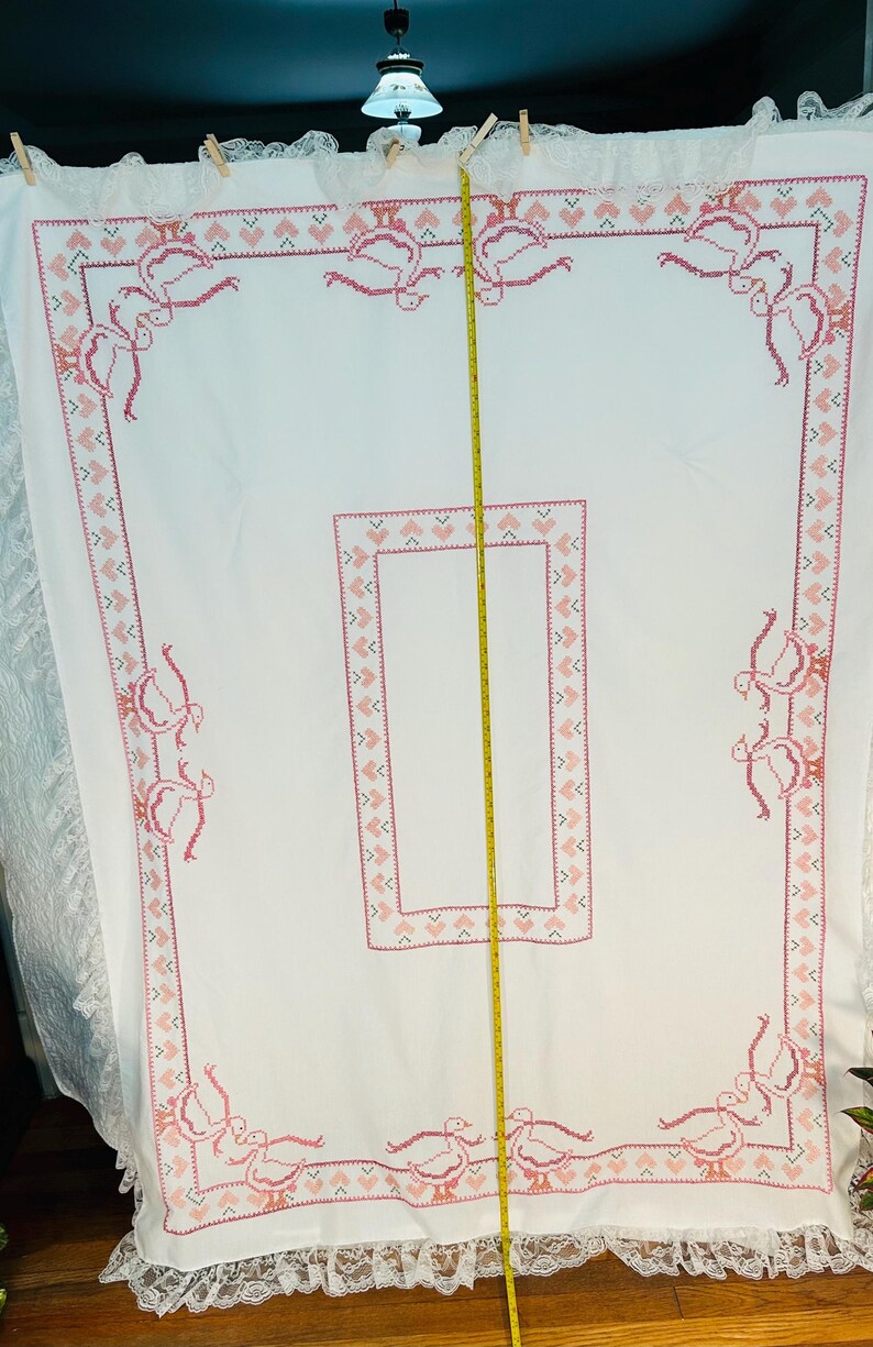 Puede incluir: Mantel blanco con un dise&ntilde;o de punto de cruz rosa. El borde presenta un patr&oacute;n repetido de corazones y p&aacute;jaros estilizados. El mantel tiene un ribete de encaje en el borde inferior. Se ve una cinta m&eacute;trica.