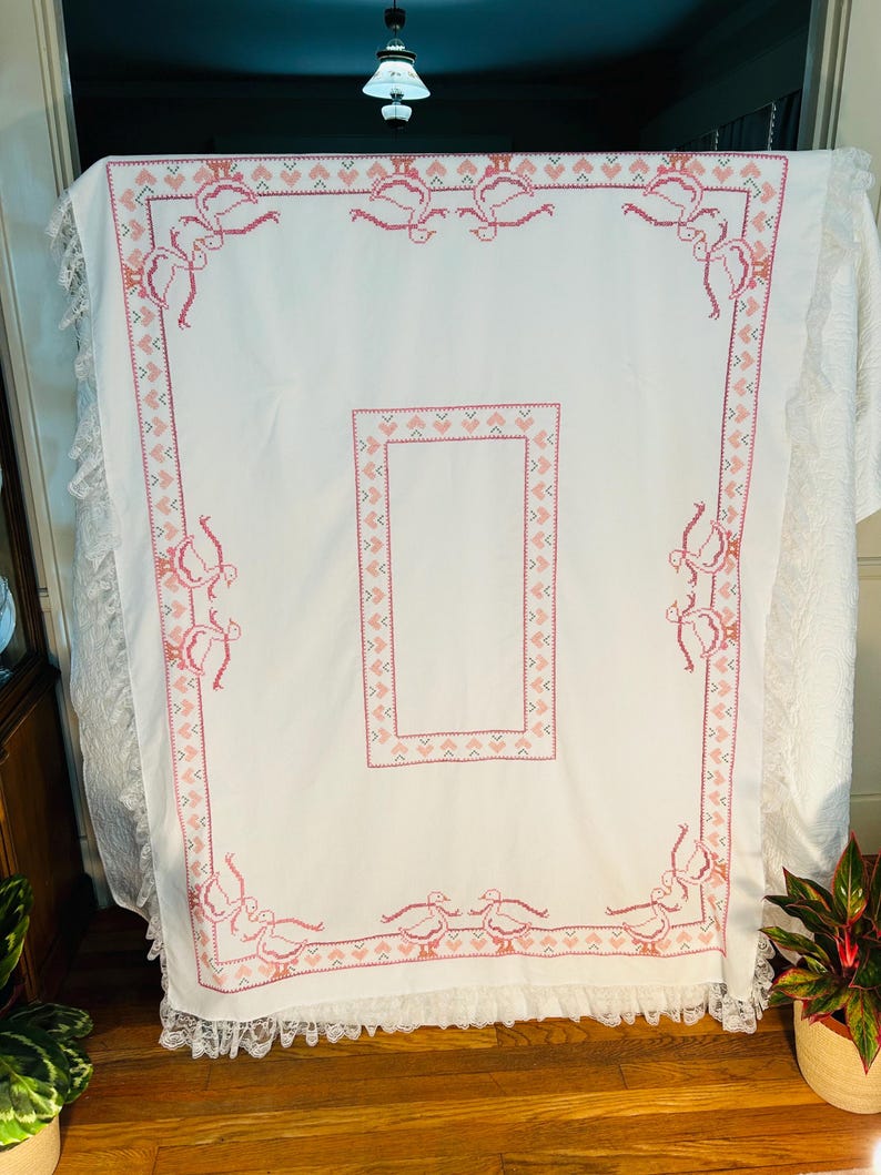 Puede incluir: Mantel blanco con un dise&ntilde;o bordado en rosa con p&aacute;jaros y patrones geom&eacute;tricos. El mantel tiene un ribete de encaje en los bordes. El dise&ntilde;o es sim&eacute;trico y cubre el borde y un &aacute;rea rectangular central.