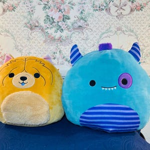 May include: Two plush toys: a yellow dog and a blue monster. The dog has a tan belly, pink ears, and a blue tongue. The monster has blue horns, a purple eye, and a striped belly. Both are round and soft.