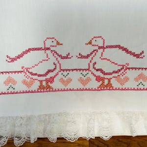 Puede incluir: Tela blanca con gansos bordados a punto de cruz en rosa y rojo, cada uno con una bufanda. Un borde de corazones rosas y detalles verdes corre debajo de los gansos. El borde inferior presenta un ribete de encaje blanco.