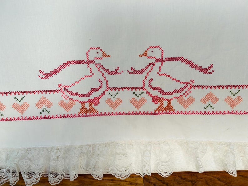 Puede incluir: Tela blanca con gansos bordados a punto de cruz en rosa y rojo, cada uno con una bufanda. Un borde de corazones rosas y detalles verdes corre debajo de los gansos. El borde inferior presenta un ribete de encaje blanco.