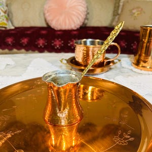 May include: A copper coffee pot with a long, textured gold-tone handle sits on a large, round copper tray featuring delicate floral engravings. In the background, a matching hammered copper mug rests on a small saucer, reflecting the warm metallic tones.