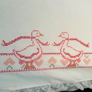 Puede incluir: Tela blanca con patos bordados a punto de cruz en rojo y rosa, cada uno con una cinta. Un borde de corazones y cruces en rosa, verde y rojo adorna el dise&ntilde;o. El borde inferior tiene un ribete de encaje.